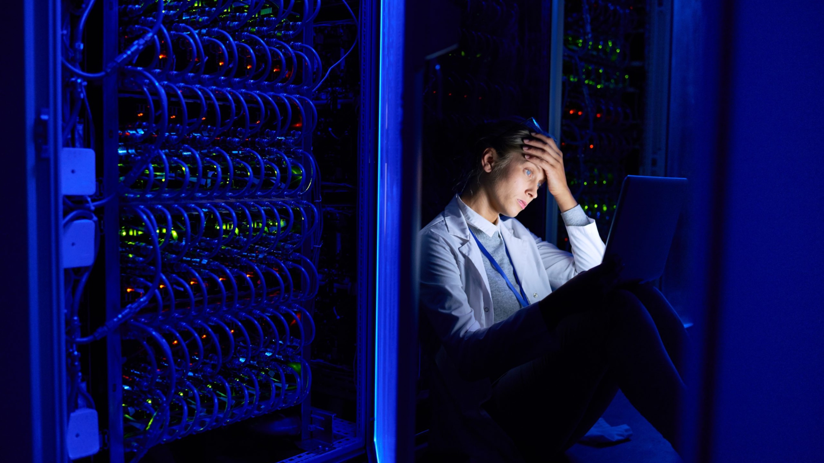 Portrait of tired young woman sitting on floor in dark server room using laptop while working with supercomputer at night